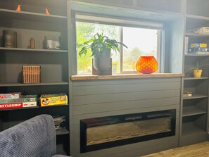 A living room with a fireplace, a window with a view of greenery, and shelves with board games. - Cozy Reno/Tahoe 3-bedroom House - Pet Friendly! (Reno)