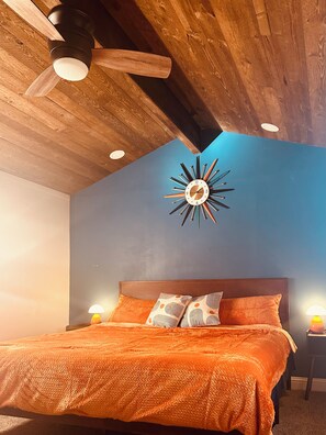A bedroom with a wooden ceiling, a bed with an orange bedspread, a wall clock, and two bedside lamps. - Cozy Reno/Tahoe 3-bedroom House - Pet Friendly! (Reno)