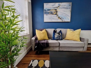 A living room with a grey sofa, yellow cushions, a blue abstract painting, and a potted plant. - Spacious, Chic & Modern Apt w/ Free Parking (Coventry)