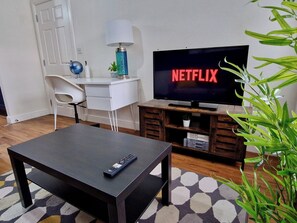 A living room with a flat-screen TV displaying 'NETFLIX', a white desk with a lamp, a chair, a coffee table, and a bookshelf. - Spacious, Chic & Modern Apt w/ Free Parking (Coventry)
