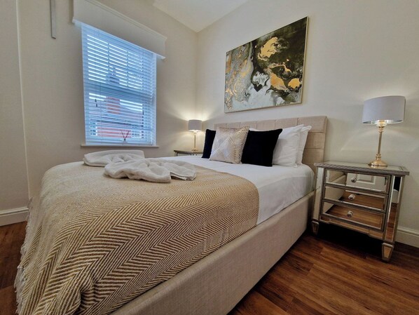 A neatly made bed with a patterned bedspread, a bedside table with a lamp, and a window with blinds. - Spacious, Chic & Modern Apt w/ Free Parking (Coventry)