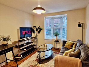 Uma sala de estar com televisão, sofá, mesa de centro e janela com persianas.