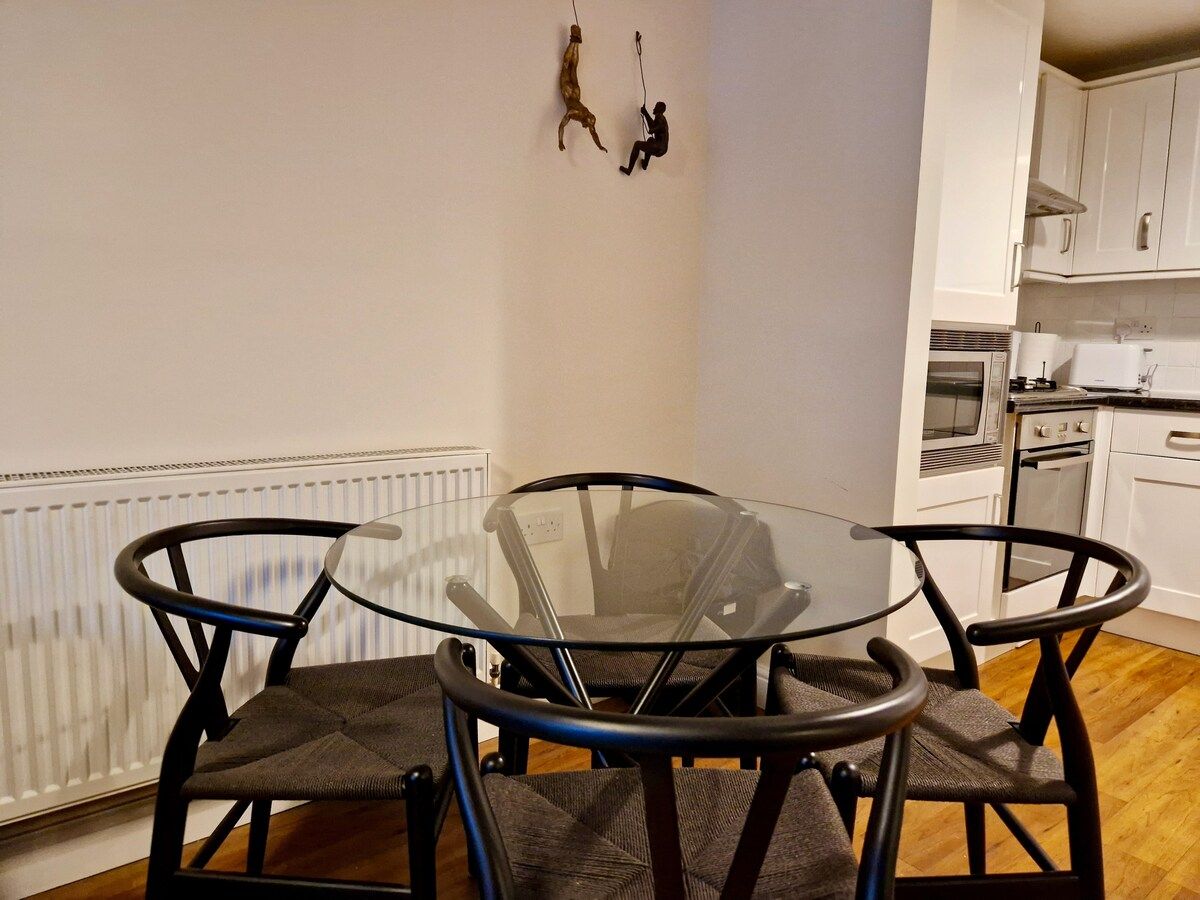 A dining area with a glass table and chairs, a white radiator, and a kitchen in the background.