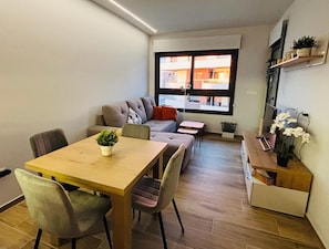 A compact living space with a dining table, sofa, and chairs, a small side table, and a window with a view of buildings. - Apartment Valentino Golf Villamartin (Orihuela)