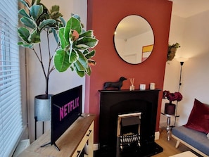 A living room with a fireplace, a Netflix sign, a potted plant, and a round mirror on the wall. - Luxurious & Cosy Home w/ Free Parking and Garden (Coventry)