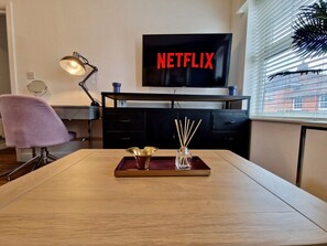 A modern living room with a wooden table, a TV displaying 'NETFLIX,' a desk lamp, and a window with blinds. - Trendy Condo Near City Centre w/ Free Parking (Coventry)