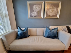 Um sofá bege com dois travesseiros azuis, um radiador e duas obras de arte florais emolduradas na parede.