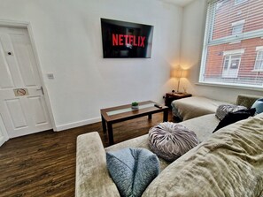 Ein Wohnzimmer mit einem an der Wand montierten Flachbildfernseher, einem kleinen Holztisch und einer Couch mit Kissen.