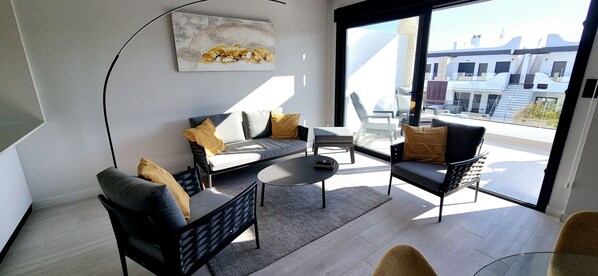 A modern living room with a sofa, two armchairs, a coffee table, and a large window offering a view of buildings. - Penthouse Innovabeach Punta Prima (Orihuela)