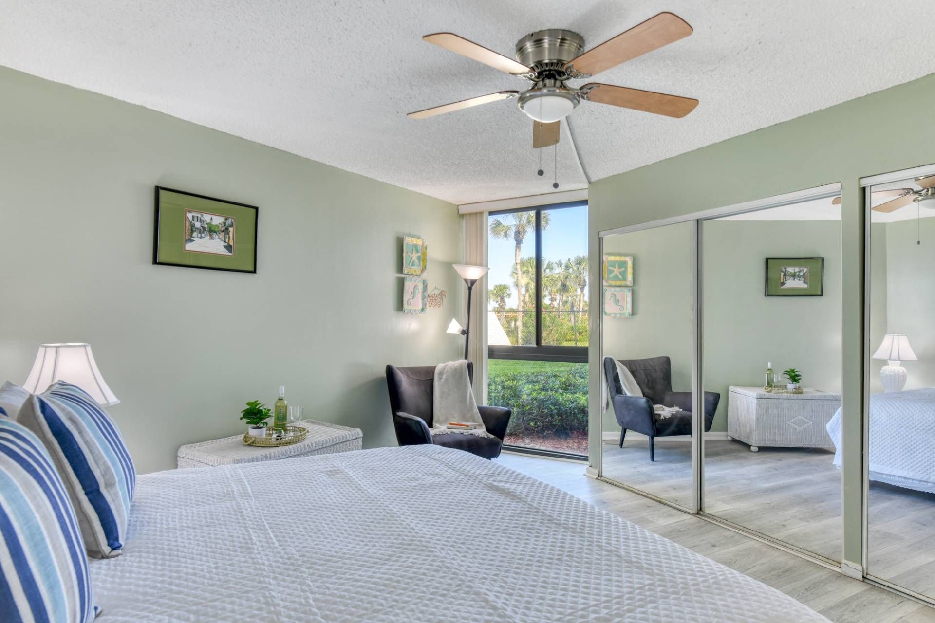 A bedroom with a bed, a ceiling fan, a nightstand, a chair, a small table, and a sliding glass door leading to a balcony.