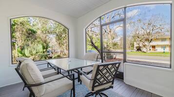 Uma sala de sol com mesa e cadeiras de vidro, grandes janelas e vista para uma área residencial.