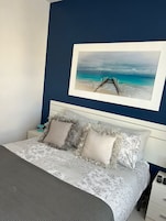 Un dormitorio con cama, almohadas, mesita de noche, cajón y un cuadro enmarcado de una playa.