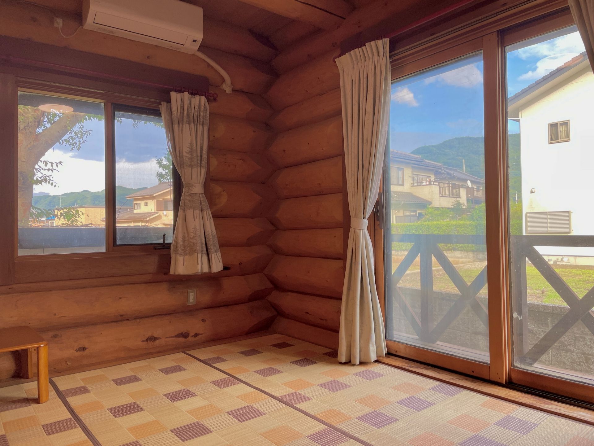 A log cabin room with a checkered floor, a window with curtains, and a view of a house and mountains.