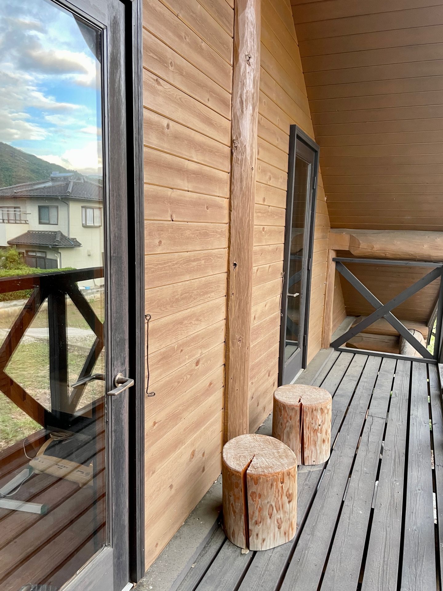 A wooden deck with a glass door leading to a room, two wooden stools, and a metal railing.