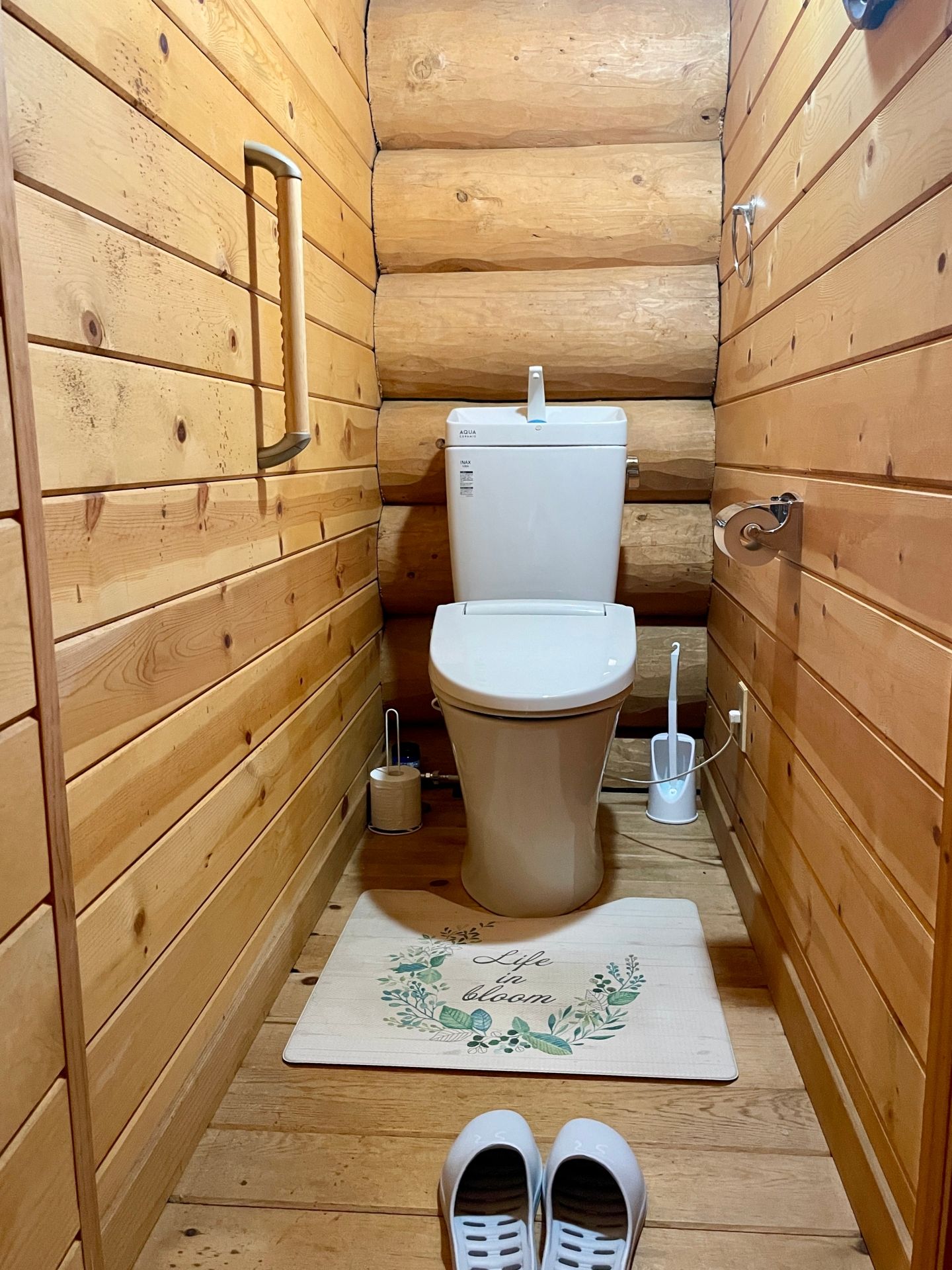 Une petite salle de bain rustique, avec des murs en bois, comprenant des toilettes, une petite poubelle, un tapis blanc à motifs floraux et une paire de pantoufles blanches posées sur le sol.