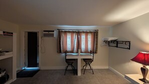 A small room with a table and two chairs, an air conditioning unit, a door, a wall-mounted shelf with a pillow, and a lamp. - Howard Motel (Windsor)