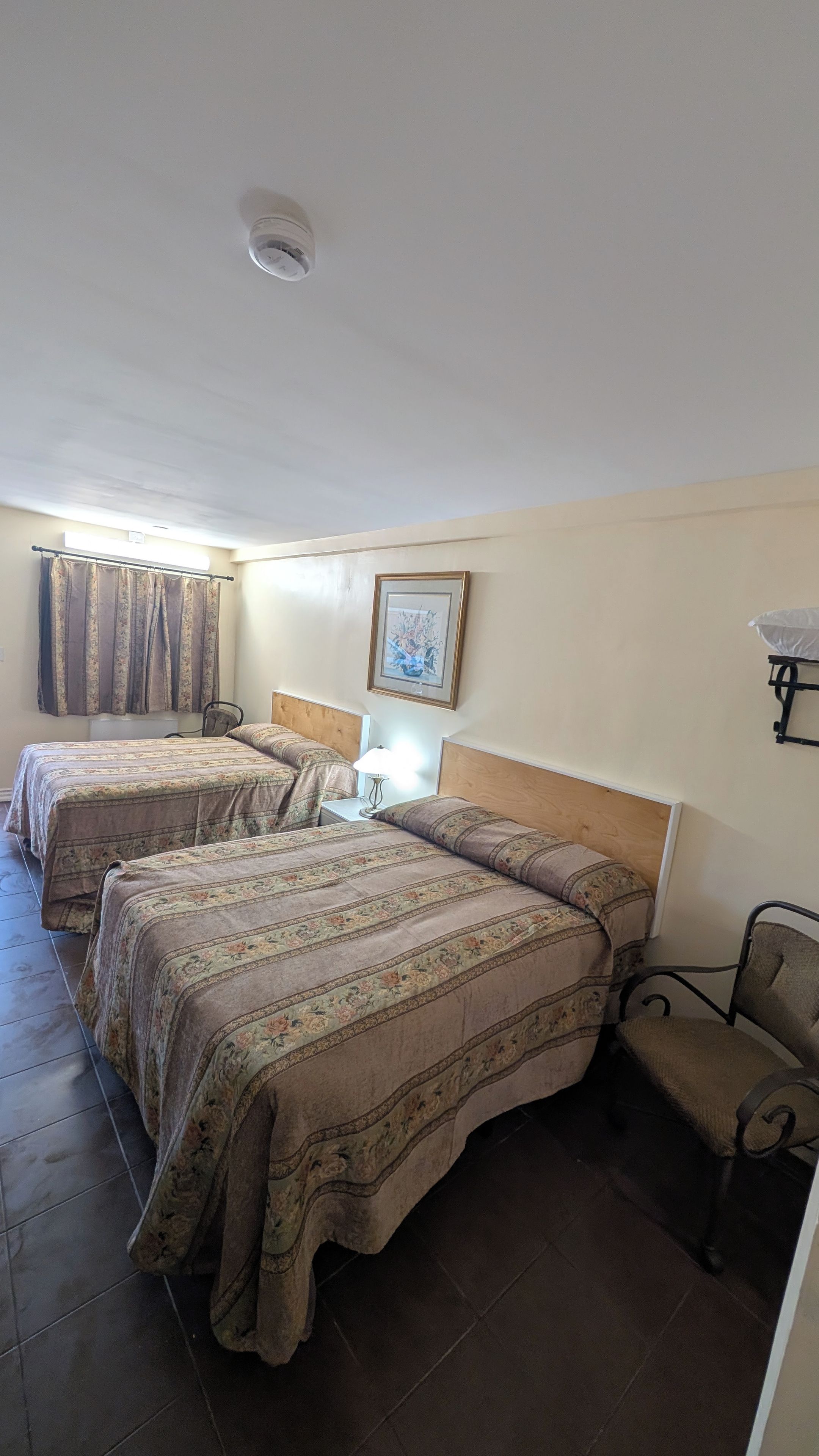 Quarto de hotel com duas camas, uma mesa de cabeceira, uma cadeira, um quadro na parede e uma luminária no teto.