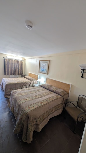 Quarto de hotel com duas camas, uma mesa de cabeceira, uma cadeira, um quadro na parede e uma luminária no teto.