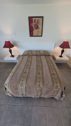 Uma cama com colcha estampada em flor, duas luminárias de cabeceira e uma obra de arte emoldurada na parede.