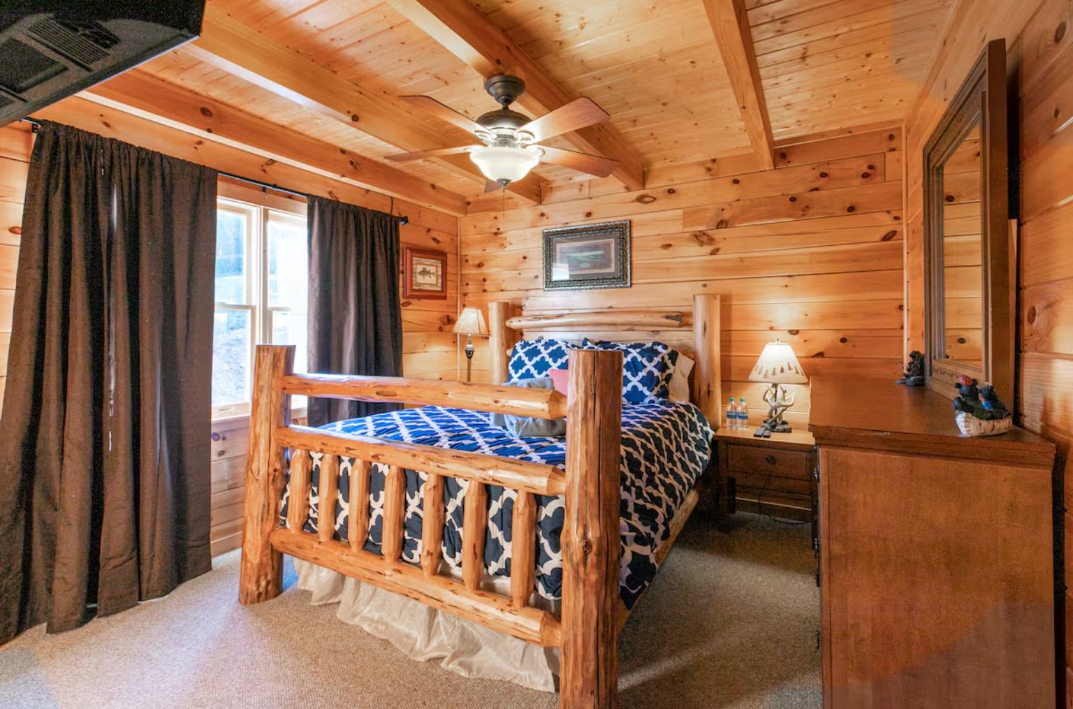 Een slaapkamer in een houten blokhut, met een bed, een dressoir, een plafondventilator en een raam met gordijnen.