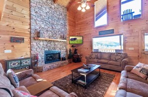 A cozy cabin interior with a stone fireplace, wooden walls, a television, and a large window. - Comfy 4-bedroom cabin with picturesque views, near Pigeon Forge and Gatlinburg (Sevierville)