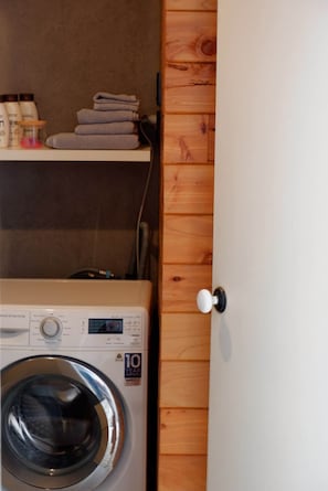Ett litet och praktiskt tvättrum med en tvättmaskin med frontmatning, en trädörr och en hylla med hopvikta handdukar och hygienartiklar.