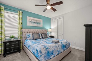 A bedroom with a tufted headboard, a bed with a blue and white patterned bedspread, a ceiling fan, a nightstand with a lamp, and a window with curtains. - 6BR Group Escape w/ Private Pool, Spa & Games (Kissimmee)