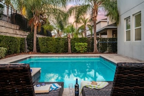 A swimming pool with a clear blue water, surrounded by palm trees and a hedge. There are two wicker lounge chairs with a bottle of wine and a book on them. - 6BR Group Escape w/ Private Pool, Spa & Games (Kissimmee)
