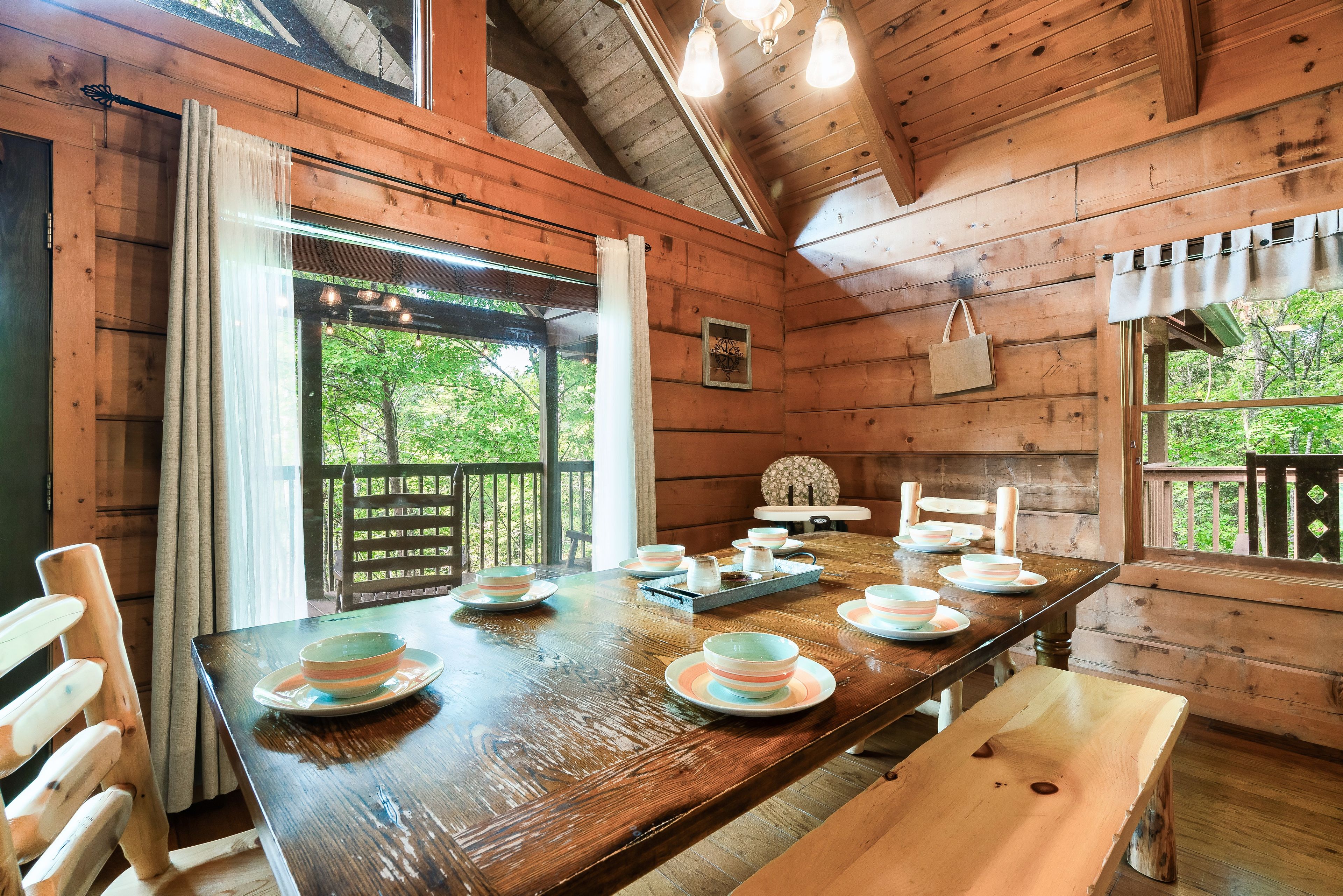 Ein Holzblockhaus-Interieur mit einem für vier Personen gedecktem Esstisch, einer Wanduhr und Blick auf viel Grün draußen.