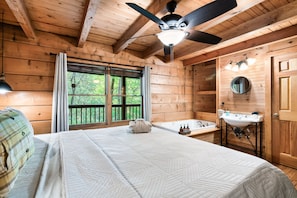 A wooden cabin bedroom with a bed, a ceiling fan, a window with curtains, a view of trees, and a bathroom with a sink and bathtub. - The ONE ! Cozy Private Cabin in a Prime Location ! (Sevierville)