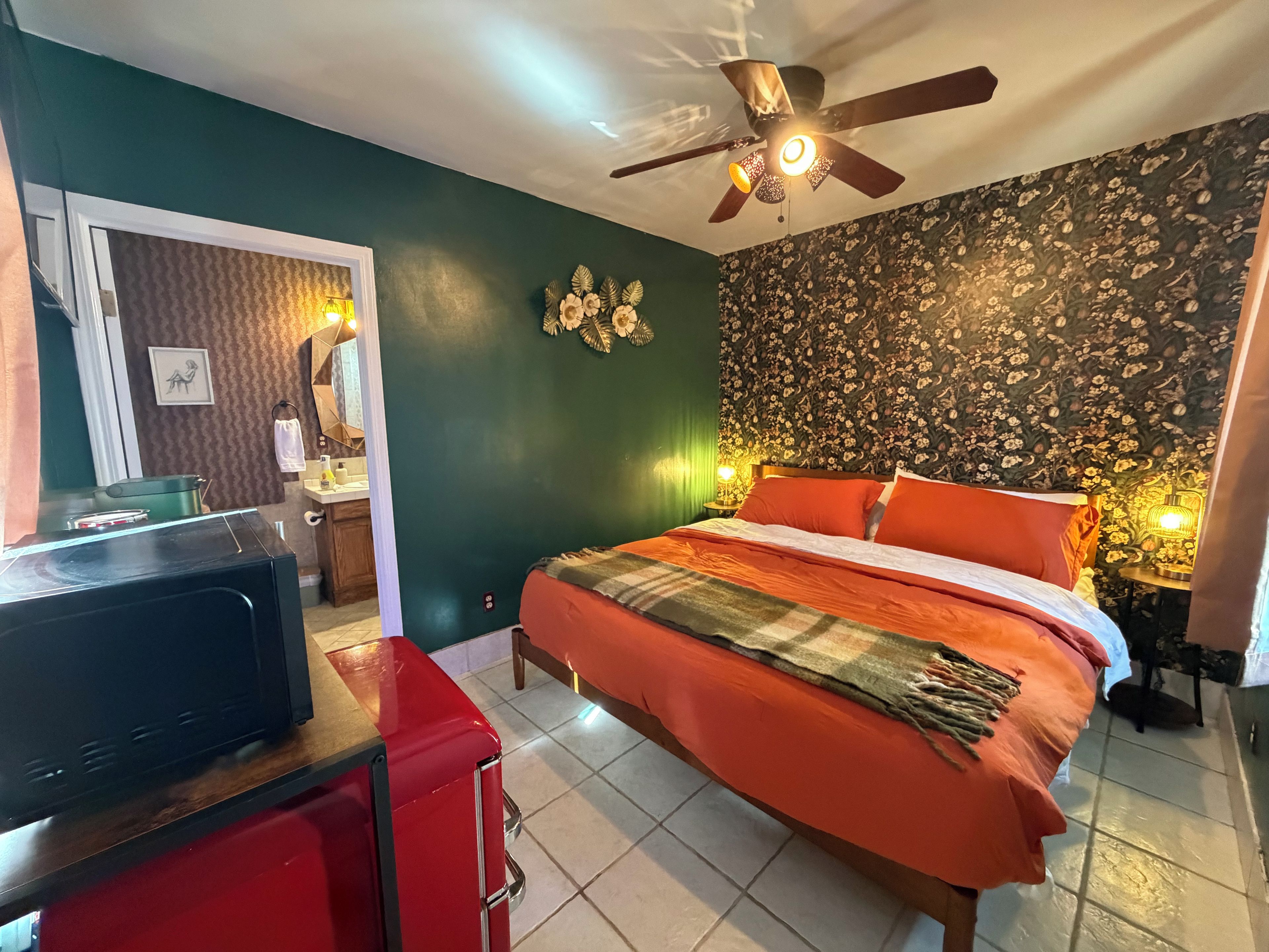 Une chambre d’hôtel avec un lit, un ventilateur de plafond, un miroir, un bureau avec une télévision et un placard rouge.