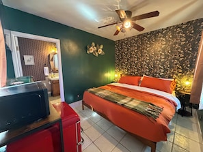 Una habitación de hotel con una cama, un ventilador de techo, un espejo, un escritorio con televisor y un armario rojo.