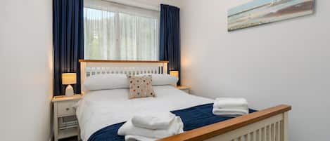 Una cama bien hecha con ropa de cama blanca y una manta azul, dos mesitas de noche con lámparas, un reloj en la pared y un cuadro sobre la cama.