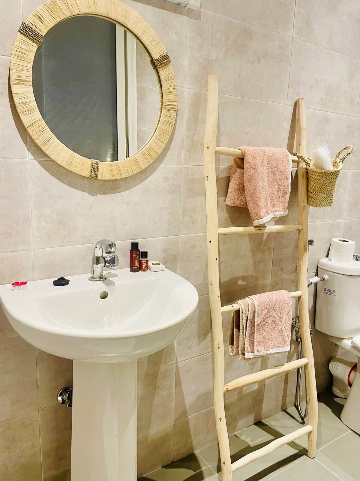 A bathroom with a round mirror, a white sink, and a wooden shelf holding towels.