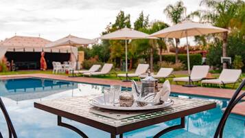 Une table placée au bord de la piscine, sur laquelle se trouve un plateau avec un service à thé, entourée de chaises longues et de parasols.