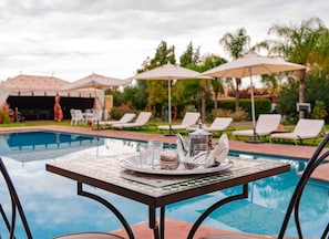 Mesa junto a la piscina con una bandeja de té, rodeada de sillones de descanso y sombrillas.