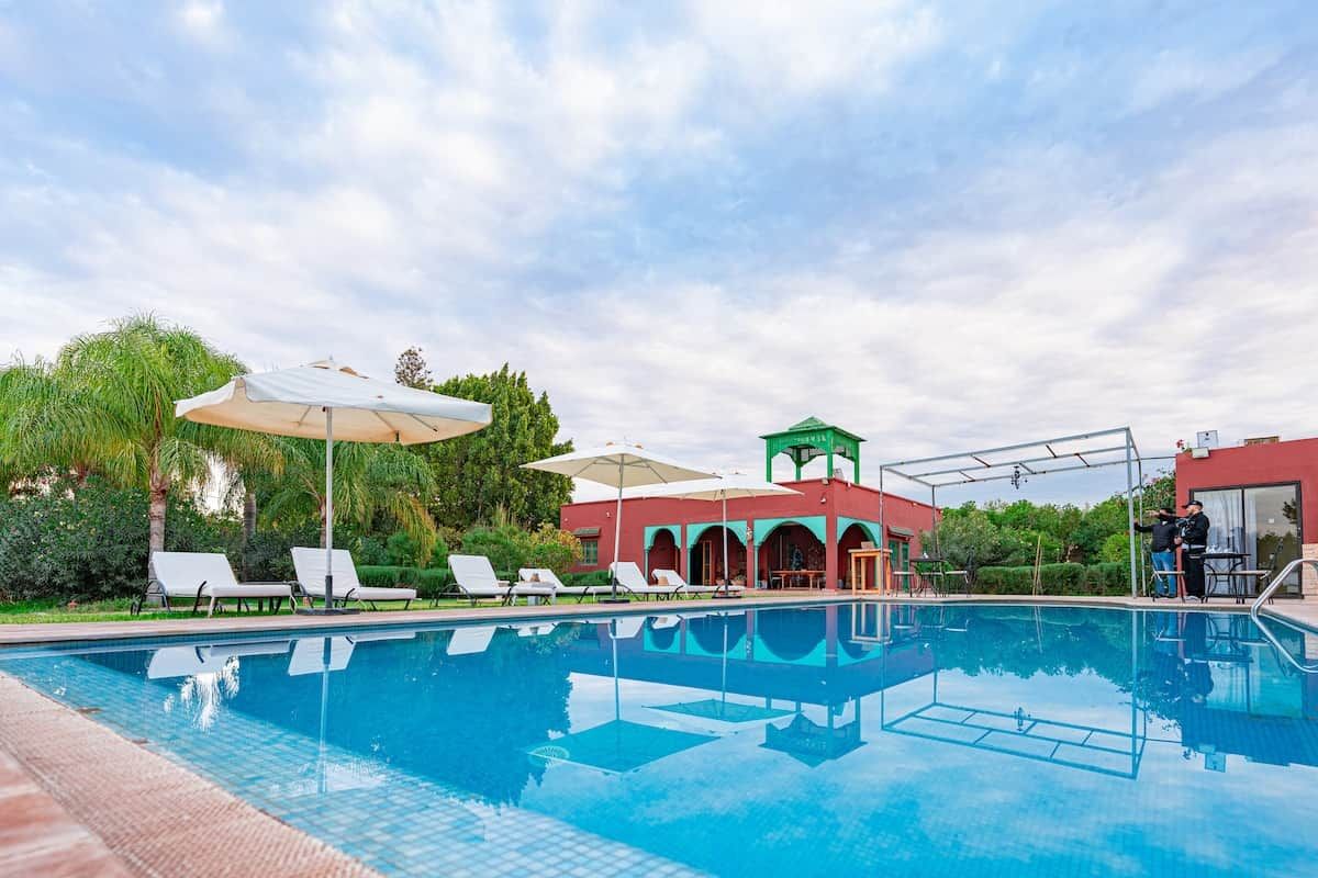 Una piscina con sillones de descanso y sombrillas, un área cubierta con techo verde y un edificio de fachada roja.
