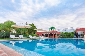 Una piscina con sillones de descanso y sombrillas, un área cubierta con techo verde y un edificio de fachada roja.