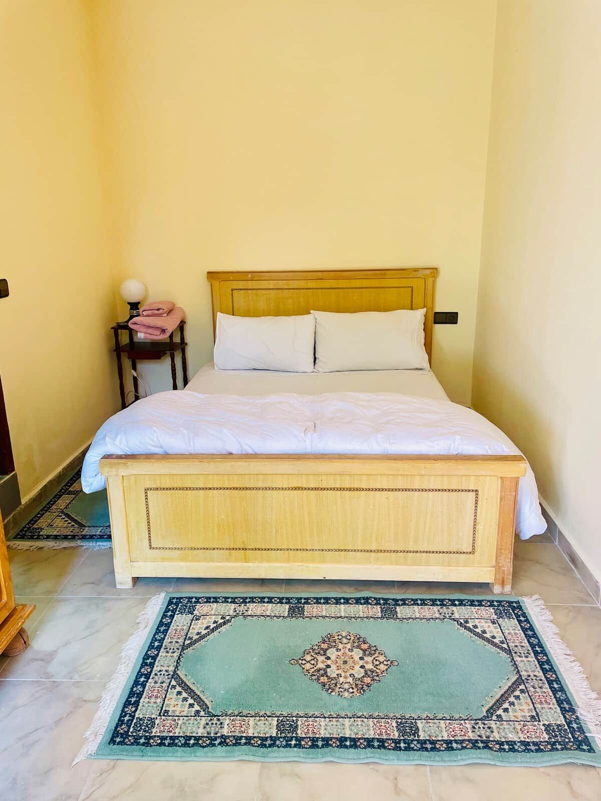 Una cama individual con cabecera de madera y ropa de cama blanca en una habitación con paredes amarillas.