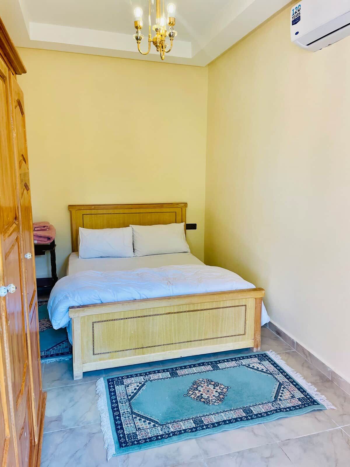 Une chambre à coucher comprenant un lit, un lustre, un appareil de climatisation, une armoire en bois et un tapis à motifs.