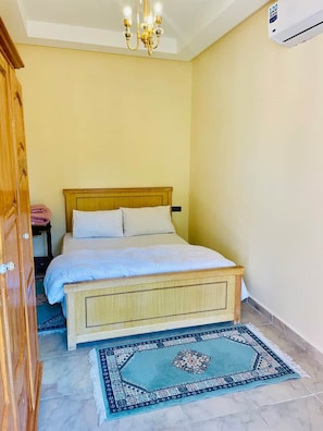 Un dormitorio con una cama, un candelabro, un equipo de aire acondicionado, un armario de madera y una alfombra estampada.