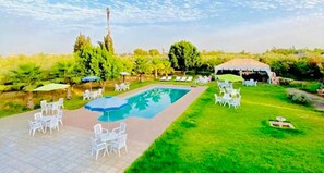 Un área de piscina con sillones de descanso y sombrillas, rodeada por un césped y árboles.