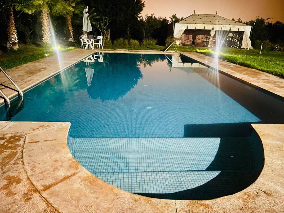 Una piscina con un borde de baldosas, un área de estar techada y un pabellón al fondo.