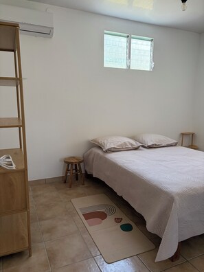 A bedroom with a bed, a wooden shelf, a small stool, and a window. - New Listing (Sainte-Luce)