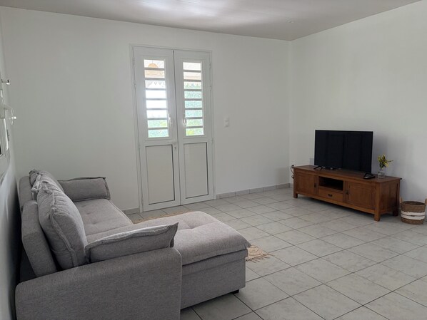 A living room with a grey sofa, a wooden TV stand, and a large window with blinds. - New Listing (Sainte-Luce)