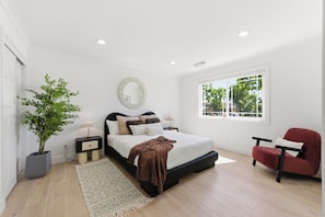 Quarto com cama, mesas de cabeceira, espelho, planta em vaso, janela e uma cadeira vermelha.