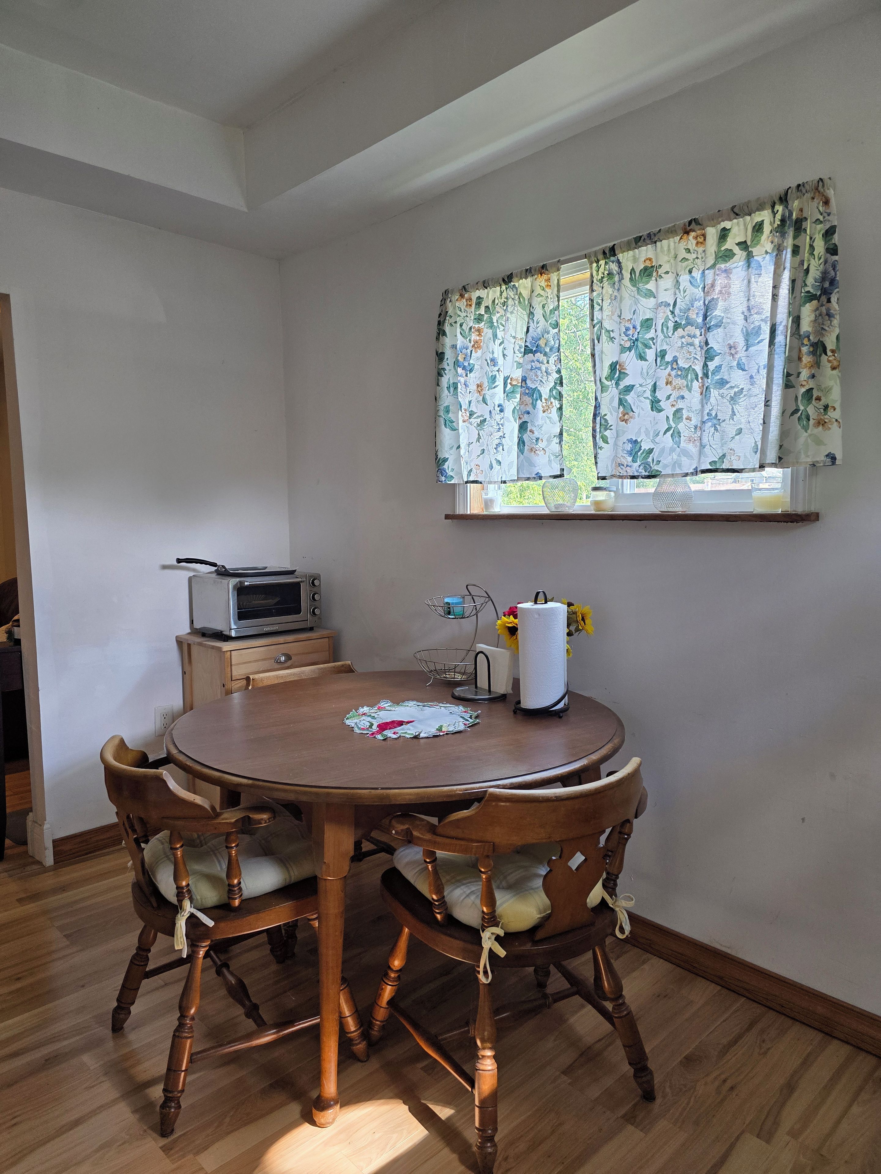 A dining area with a round wooden table, four chairs, a small kitchen unit, and a window with floral curtains.