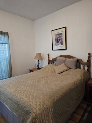 A bedroom with a large bed, a nightstand with a lamp, a framed picture on the wall, and a window with blinds. - Carico Comfortable Family Home Near Champaign (Tuscola)