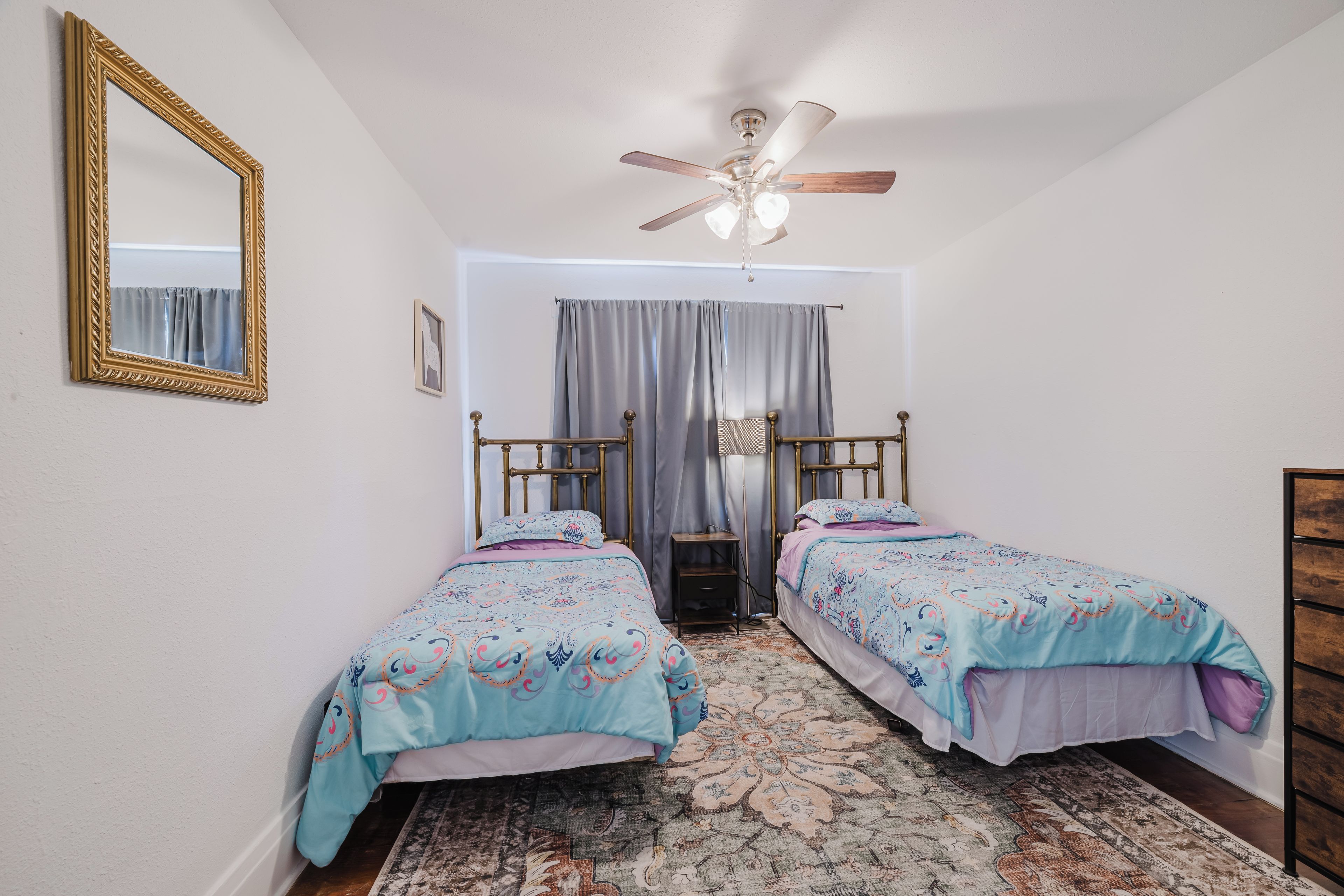 Une chambre avec deux lits simples, un ventilateur de plafond, un grand miroir et un tapis à motifs.