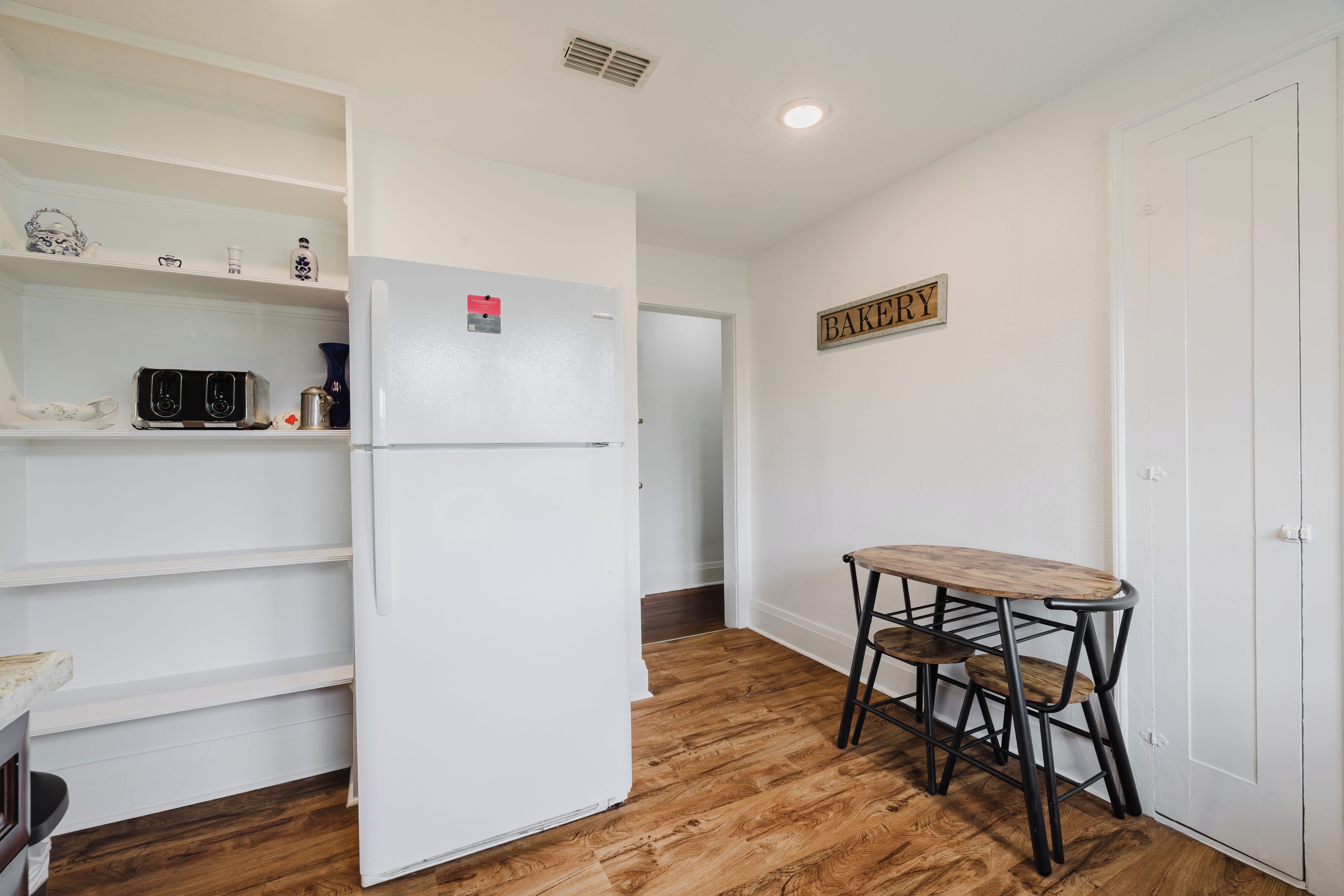 A compact kitchen with a white refrigerator, a small table with two chairs, and a 'BAKERY' sign on the wall.